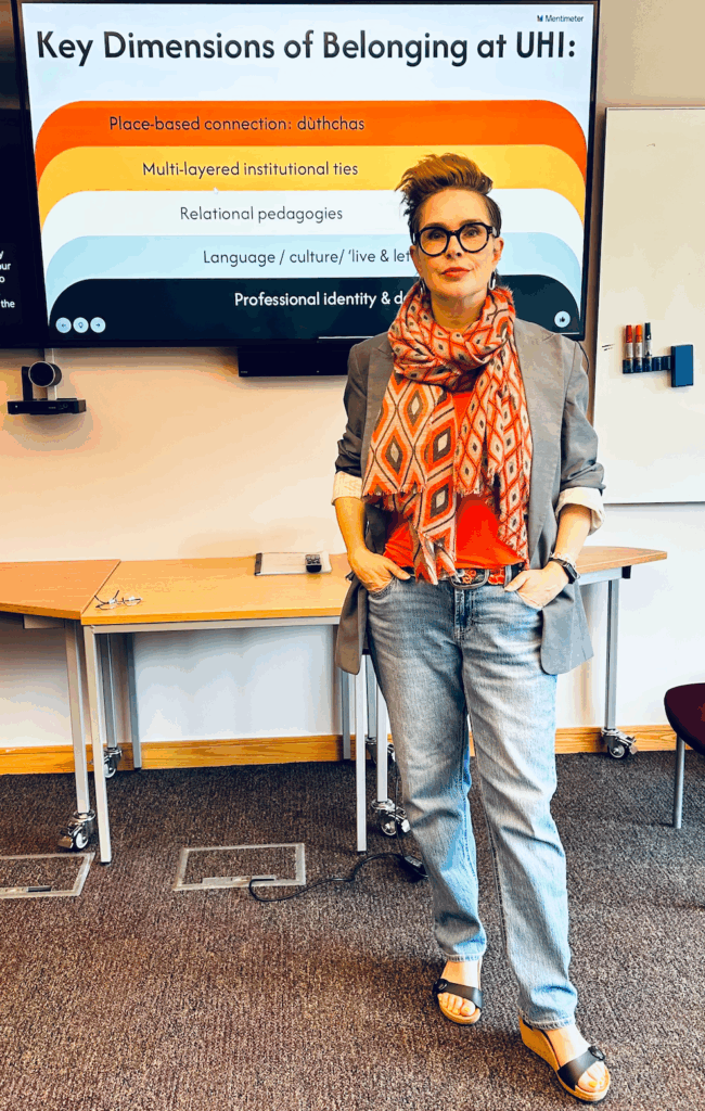A white person with short dyed reddish hair and glasses, in jeans, a blue blazer, and a femme scarf stands in front of a slide reading "Key Dimensions of Belonging at UHI" with rainbow-ish colours below and info on each colour. The dimensions listed are: Place-based connection, Multi-layered institutional ties, Relational pedagogies, Language/culture/"live and let live" ethos (not fully readable) and Professional identity and development (again, not fully readable).